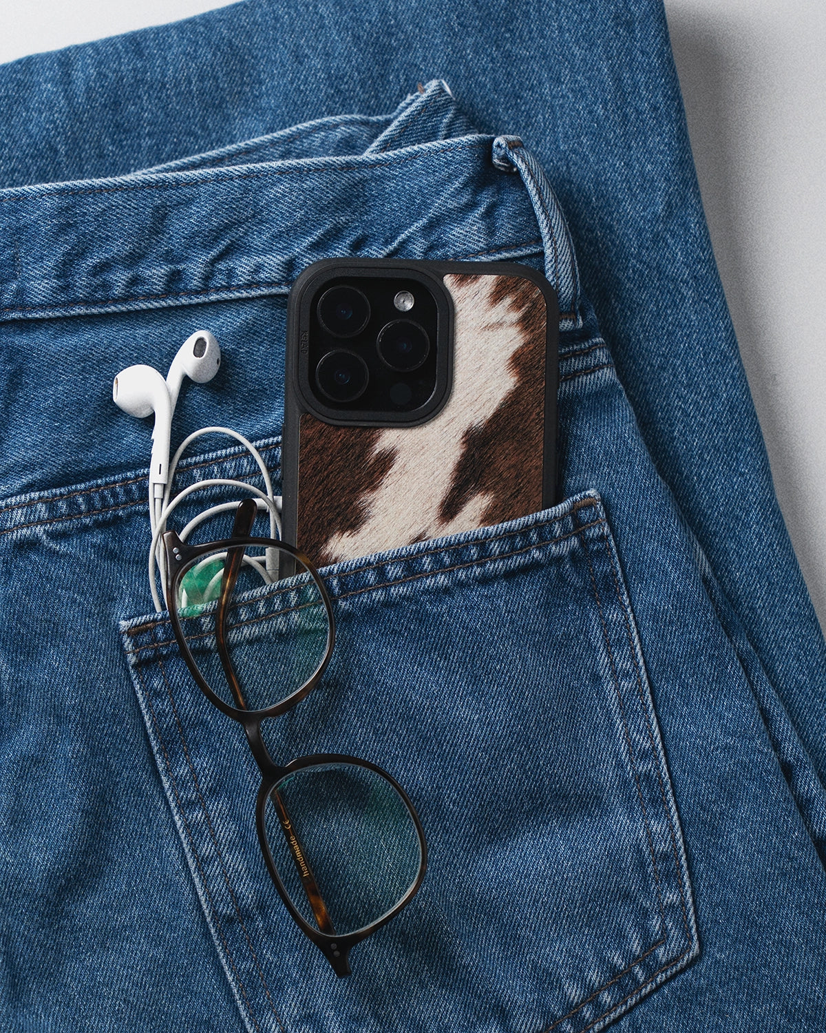 Phone case with cow print, earphones, and glasses in a blue denim pocket.
