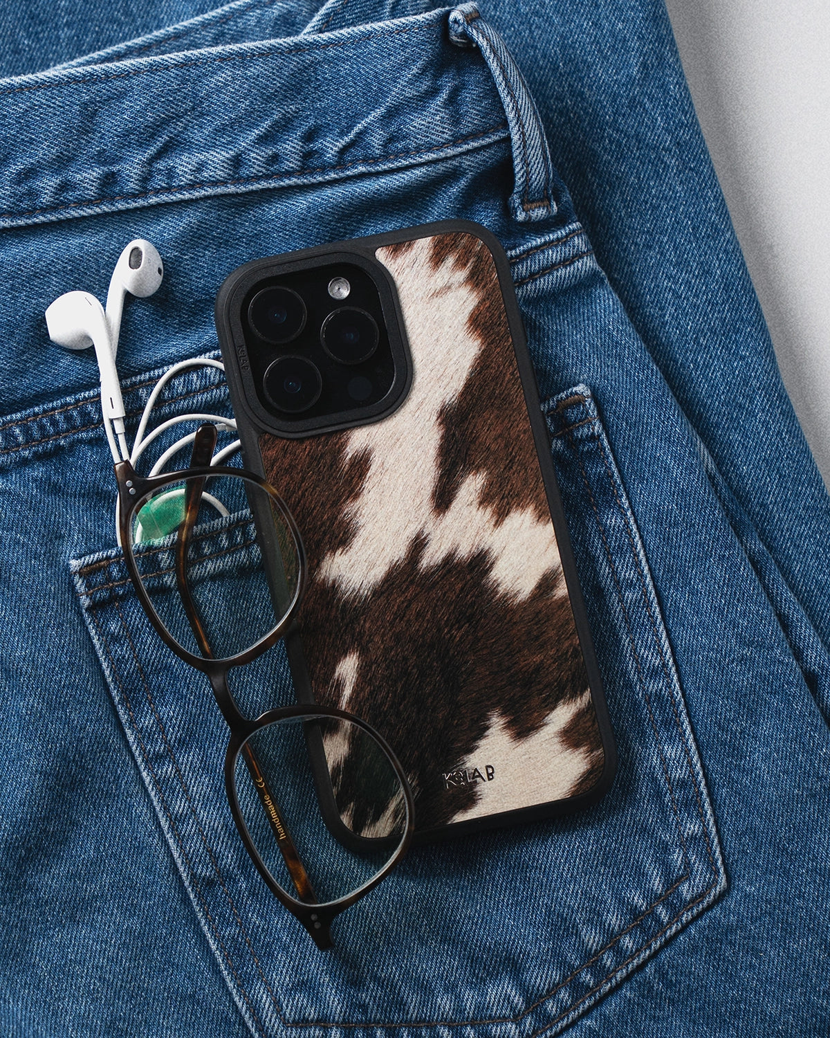 KELAB iPhone with cow print case, glasses, and earbuds on a blue denim background