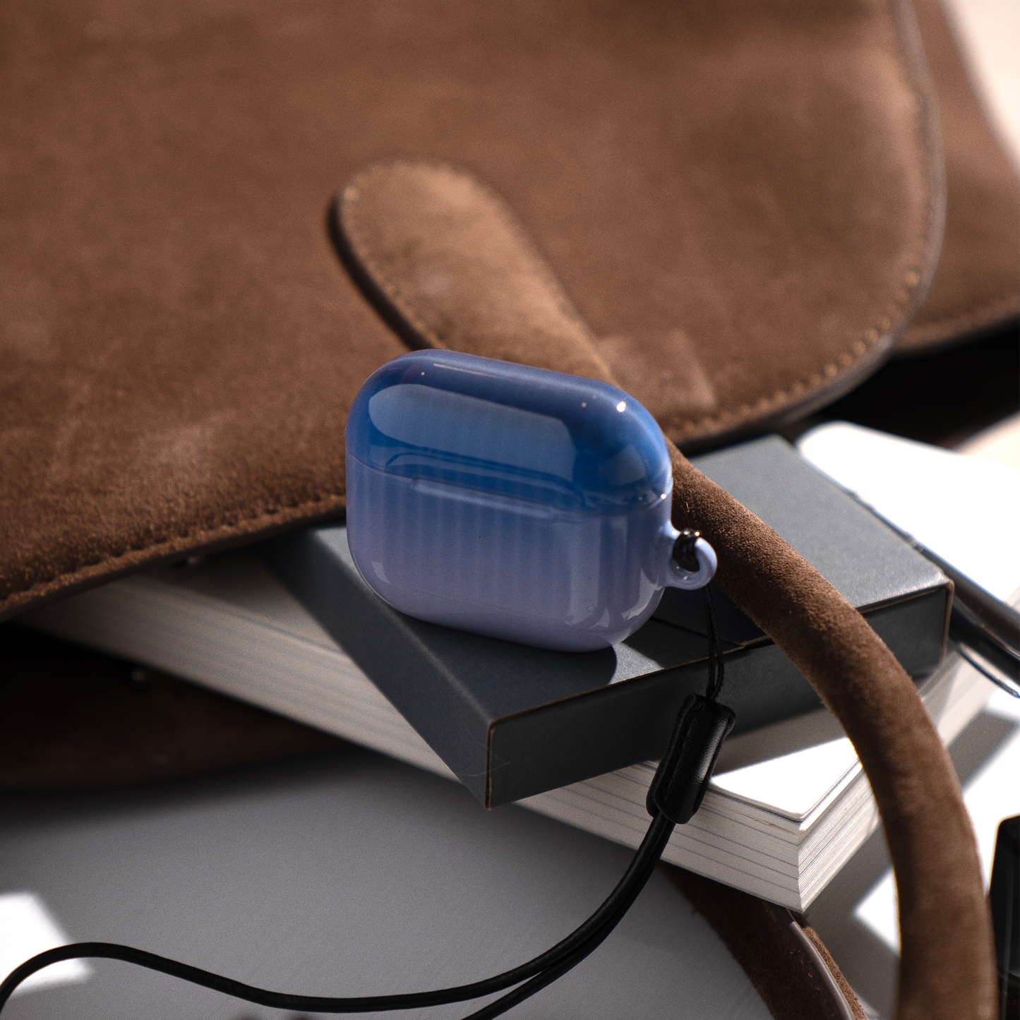 Twilight Cascade Blue AirPods Case from KELAB resting on books beside a suede bag, highlighting its stylish ombre design and portability.