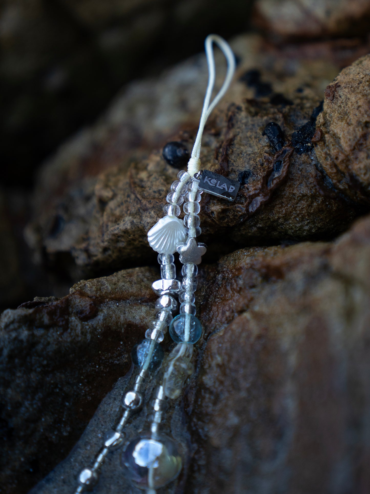 Crystal Sea Star Phone Charm with beaded shells, chrome stars, and glass beads on a white strap, displayed on coastal rocks. Ideal beach-themed phone accessory.