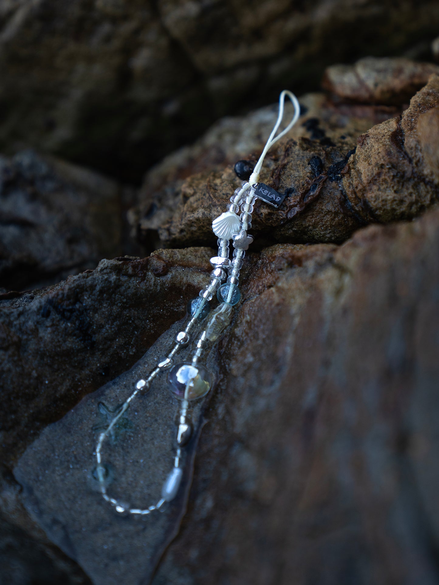 Crystal Sea Star Phone Charm with beaded shells, stars, and chrome accents on a rocky beach background. Ideal as a phone strap or keychain accessory.