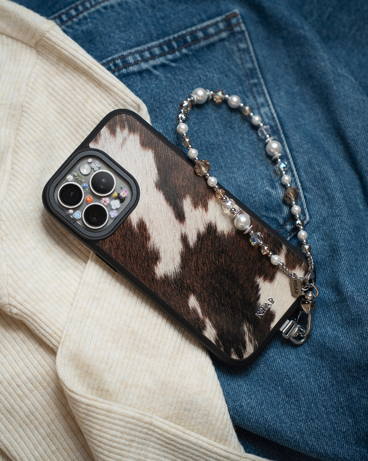 KELAB iPhone case with cow print and beaded strap on a blue and beige background